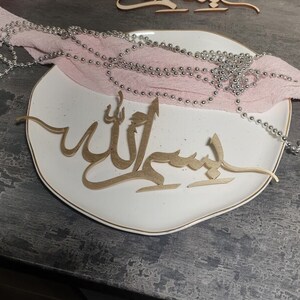 Op de afbeelding: Een wit bord met een gouden rand en een gouden houten uitsnijding van het Arabische woord "Allah" erop. Een roze stof met zilveren kralen is over het bord gelegd.