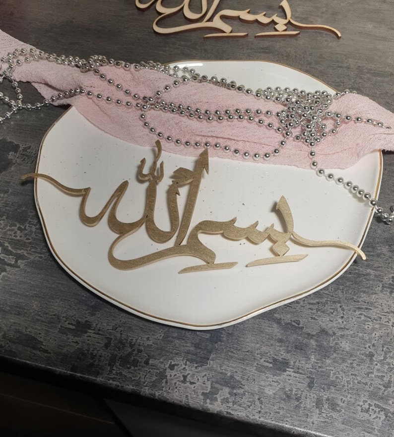 Op de afbeelding: Een wit bord met een gouden rand en een gouden houten uitsnijding van het Arabische woord "Allah" erop. Een roze stof met zilveren kralen is over het bord gelegd.