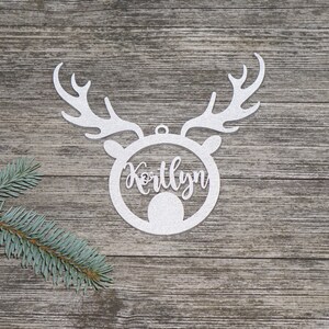 May include: A white, glittery reindeer head ornament with the name "Kortlyn" in a circular frame. The ornament has antlers and a small loop for hanging. A sprig of evergreen is in the lower left corner, set against a wood-grain background.