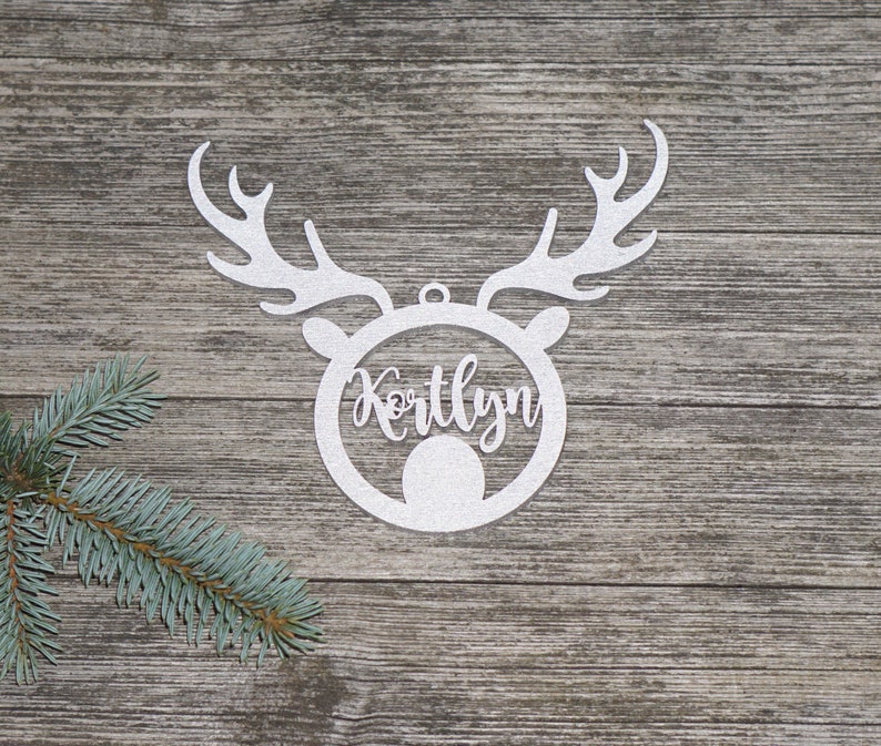 May include: A white, glittery reindeer head ornament with the name "Kortlyn" in a circular frame. The ornament has antlers and a small loop for hanging. A sprig of evergreen is in the lower left corner, set against a wood-grain background.