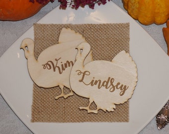 Puede incluir: Dos tarjetas de lugar de madera con forma de pavo, con los nombres "Kim" y "Lindsey" grabados. Se colocan sobre un cuadrado de arpillera en un plato blanco, con calabazas y follaje otoñal en el fondo.