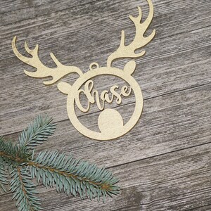 May include: A gold-colored wooden Christmas ornament in the shape of a reindeer head with antlers. The name "Chase" is cut out in cursive within the reindeer's face. A sprig of evergreen is in the lower left corner.