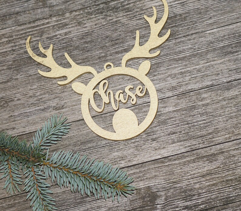 May include: A gold-colored wooden Christmas ornament in the shape of a reindeer head with antlers. The name "Chase" is cut out in cursive within the reindeer's face. A sprig of evergreen is in the lower left corner.
