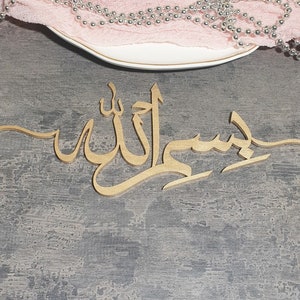 Op de afbeelding: Een gouden houten bord met de Arabische zin "Bismillah" in kalligrafie. Het bord ligt op een grijs, getextureerd oppervlak.