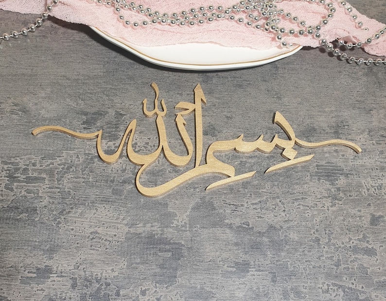 Op de afbeelding: Een gouden houten bord met de Arabische zin "Bismillah" in kalligrafie. Het bord ligt op een grijs, getextureerd oppervlak.