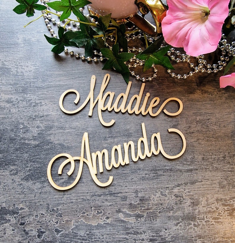 May include: Two wooden signs with the names Maddie and Amanda written in cursive script. The signs are on a gray wooden surface.