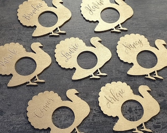 Puede incluir: Tarjetas de mesa con forma de pavo de madera clara, grabadas con nombres como "Brendan" y "Kevin" en escritura cursiva. Cada tarjeta tiene una abertura circular. Perfectas para Acción de Gracias.
