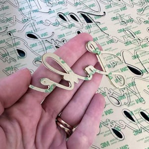 May include: A gold-colored, cursive-style word "love" cut-out, held in a hand. The word is adhered to a backing with green adhesive. The background features a repeating pattern of the same word cut-out.