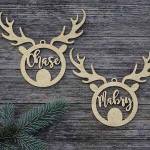 May include: Two wooden reindeer head ornaments with the names "Chase" and "Mabry" cut out in a cursive font. The ornaments have antlers and a circular body. A sprig of evergreen is in the lower left corner.