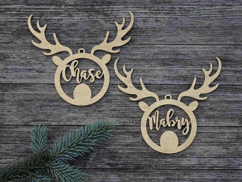May include: Two wooden reindeer head ornaments with the names "Chase" and "Mabry" cut out in a cursive font. The ornaments have antlers and a circular body. A sprig of evergreen is in the lower left corner.