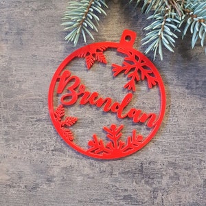 May include: Red Christmas ornament with the name "Brunolan" cut out in the center. The ornament is decorated with snowflakes and leaves.