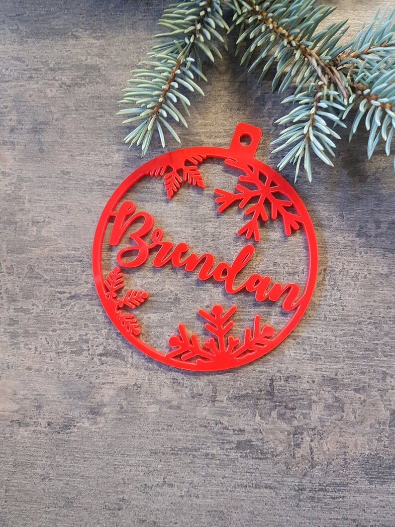 May include: Red Christmas ornament with the name "Brunolan" cut out in the center. The ornament is decorated with snowflakes and leaves.