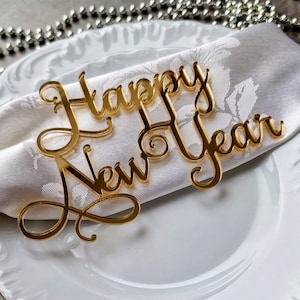 Op de afbeelding: Een goudkleurige tafeldecoratie met de tekst "Happy New Year" in sierlijke letters, rustend op een witte servet, geplaatst op een wit bord. Het elegante schrift reflecteert en voegt een feestelijke toets toe. Zilveren kralen zijn zichtbaar op de achtergrond.