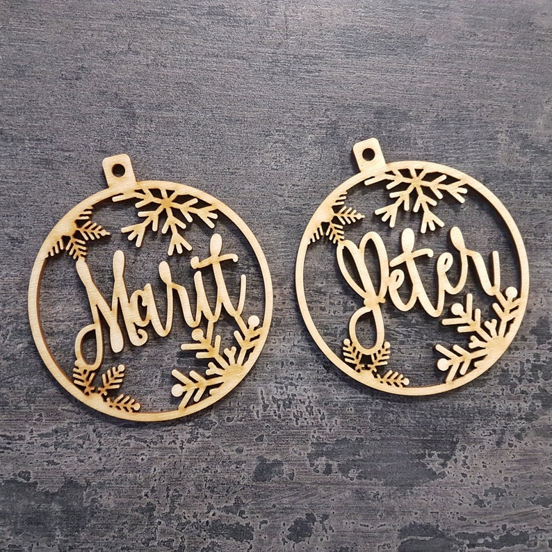 May include: Two wooden Christmas baubles. Each bauble is a circle with a snowflake design and a name cut out. One says "Marit" and the other says "Peter". Both have a small hole at the top for hanging.