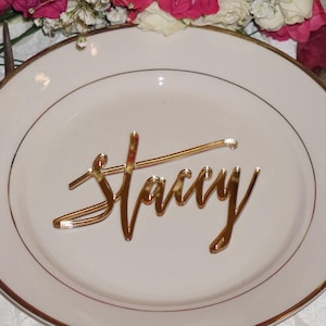May include: Gold mirror acrylic place card with the name "Stacy" written in a cursive font.