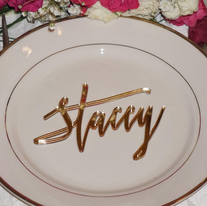 May include: Gold mirror acrylic place card with the name "Stacy" written in a cursive font.