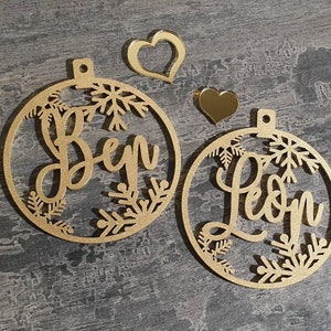 May include: Two gold-colored wooden ornaments with snowflake designs. The ornaments are personalized with the names "Ben" and "Leon".