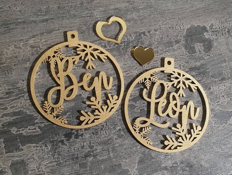 May include: Two gold-colored wooden ornaments with snowflake designs. The ornaments are personalized with the names "Ben" and "Leon".