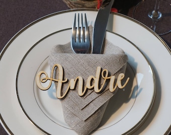 Puede incluir: Una tarjeta de lugar de madera con el nombre "André" escrito en cursiva. La tarjeta de lugar está colocada sobre una servilleta de lino blanco con un tenedor y un cuchillo.