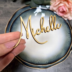 Può includere: Un segnaposto in legno con il nome "Michelle" scritto in corsivo. Il segnaposto è su un piatto blu e bianco.