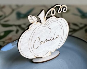Könnte beinhalten: Ein Platzkartenhalter in Kürbisform aus Holz mit dem Namen "Camila" darauf. Die Platzkarte ist hell gefärbt mit einem dunkelbraunen Sockel und Stiel. Der Kürbis hat ein Blatt und eine lockige Ranke.