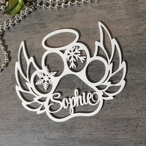 Op de afbeelding: Wit houten poot afdruk ornament met engelenvleugels, een halo en sneeuwvlokken. De naam "Sophie" is geschreven in de poot afdruk.