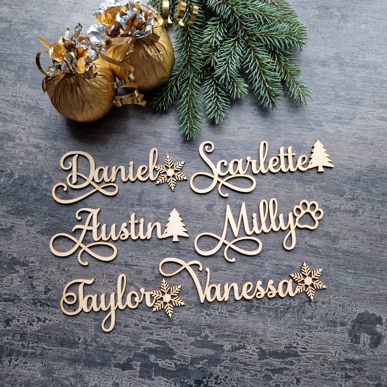 May include: Wooden Christmas ornaments with names "Daniel", "Scarlette", "Austin", "Milly", "Taylor", and "Vanessa" with snowflake and tree designs.