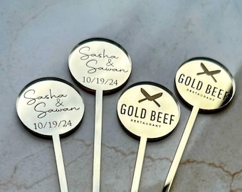 May include: Three gold-colored round acrylic signs with sticks attached. The signs have different text. One sign says "Sasha & Sawan 10/19/24". Another sign says "Gold Beef Restaurant" with a crossed knife and fork design. The third sign also says "Gold Beef Restaurant" with a crossed knife and fork design.