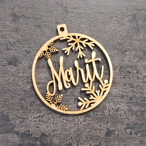 May include: A wooden Christmas bauble with the name "Marit" cut out in a cursive font. The bauble is round with snowflake and leaf cutouts around the edges. It has a small hole at the top for hanging.