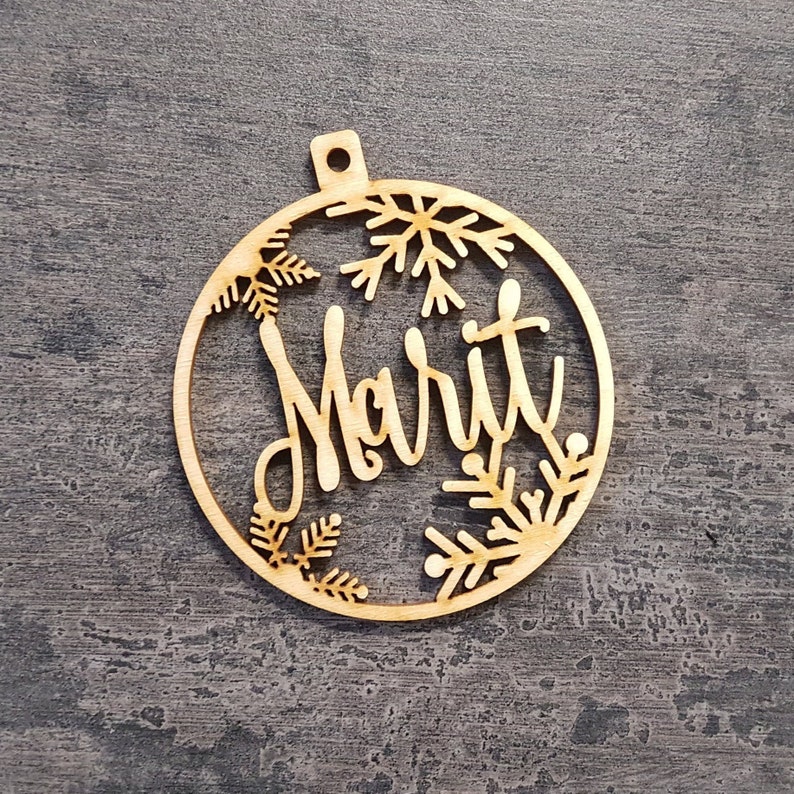May include: A wooden Christmas bauble with the name "Marit" cut out in a cursive font. The bauble is round with snowflake and leaf cutouts around the edges. It has a small hole at the top for hanging.