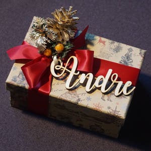 Puede incluir: Una caja de regalo envuelta en papel marr&oacute;n con una cinta roja y un adorno de pi&ntilde;a. La caja est&aacute; decorada con una etiqueta de nombre de madera blanca que dice "Andr&eacute;".