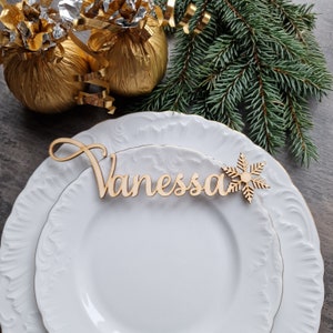 May include: A white plate with a wooden name card that says "Vanessa" and a snowflake cutout. The plate is on a wooden table with gold wrapping paper and a green pine branch in the background.