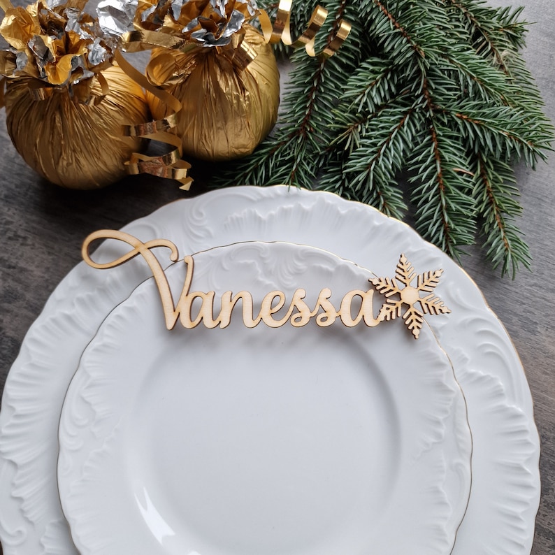 May include: A white plate with a wooden name card that says "Vanessa" and a snowflake cutout. The plate is on a wooden table with gold wrapping paper and a green pine branch in the background.