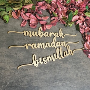 Op de afbeelding: Drie gouden houten borden met de woorden "mubarak", "ramadan" en "bismillah" geschreven in een cursief schrift. De borden liggen op een grijze houten achtergrond met roze en groene bloemblaadjes die eromheen zijn verspreid.