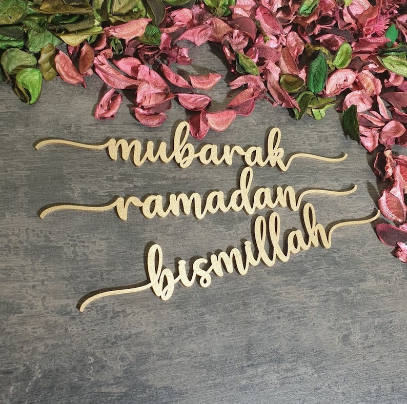 Op de afbeelding: Drie gouden houten borden met de woorden "mubarak", "ramadan" en "bismillah" geschreven in een cursief schrift. De borden liggen op een grijze houten achtergrond met roze en groene bloemblaadjes die eromheen zijn verspreid.