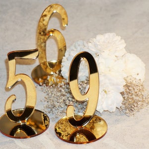 Puede incluir: Tres números de mesa de acrílico dorado espejado, 5, 6 y 9, sobre pequeñas bases redondas. Los números están sobre una superficie blanca con flores blancas y flores secas.