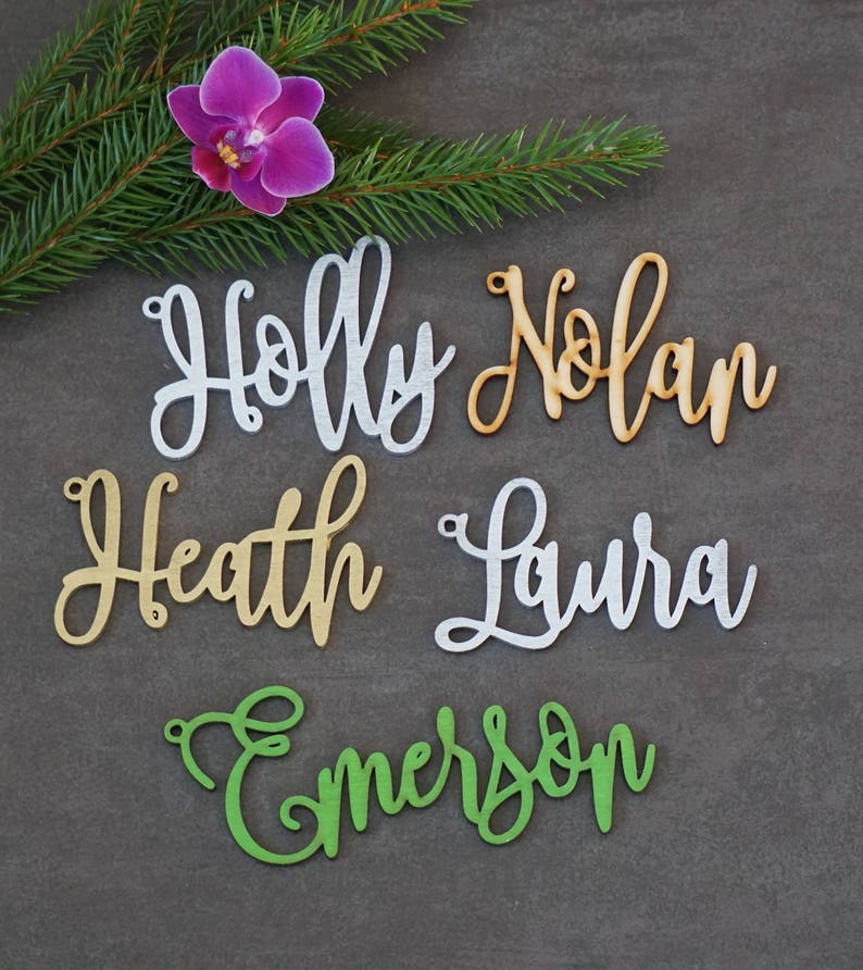 May include: Five wooden name tags with cursive lettering. The names are Holly, Nolan, Heath, Laura, and Emerson. The names are in different colors: silver, brown, gold, silver, and green.