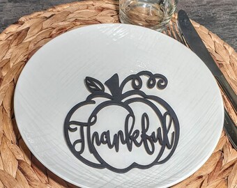 Könnte beinhalten: Eine schwarze, kürbisförmige Tischkarte mit dem Wort "Thankful" in Schreibschrift, platziert auf einem weißen Teller. Der Teller steht auf einem gewebten Platzset, mit einem Glas und Besteck im Hintergrund. Eine herbstliche Tischdekoration.