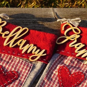 May include: Two red and gray Christmas stockings with personalized wooden name tags. The stockings have a red heart sewn on the front. The name tags read "Bellamy" and "Grammy".