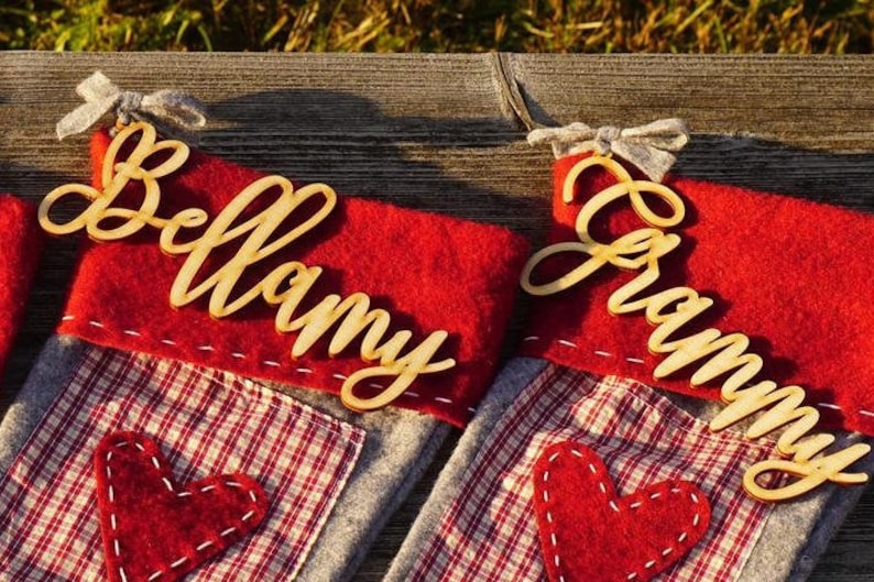 May include: Two red and gray Christmas stockings with personalized wooden name tags. The stockings have a red heart sewn on the front. The name tags read "Bellamy" and "Grammy".