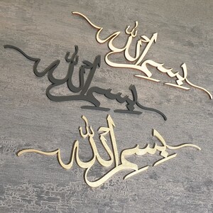 Op de afbeelding: Drie houten wanddecoraties met de Arabische zin "Bismillah" in zwarte en gouden kalligrafie. De zin wordt drie keer herhaald, met het bovenste stuk in zwart, het middelste stuk in zwart en het onderste stuk in goud.