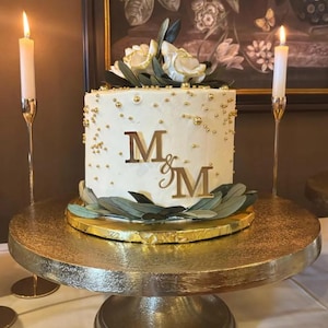 May include: A white iced cake decorated with gold sprinkles, flowers, and olive leaves. The cake has the letters "M & M" in gold. It sits on a gold cake stand, with two lit candles in the background.