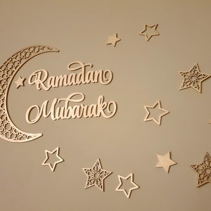 May include: A wooden crescent moon with intricate cutouts and the words "Ramadan Mubarak" in cursive script. The moon is surrounded by 10 wooden stars, some with intricate cutouts and some with simple designs.
