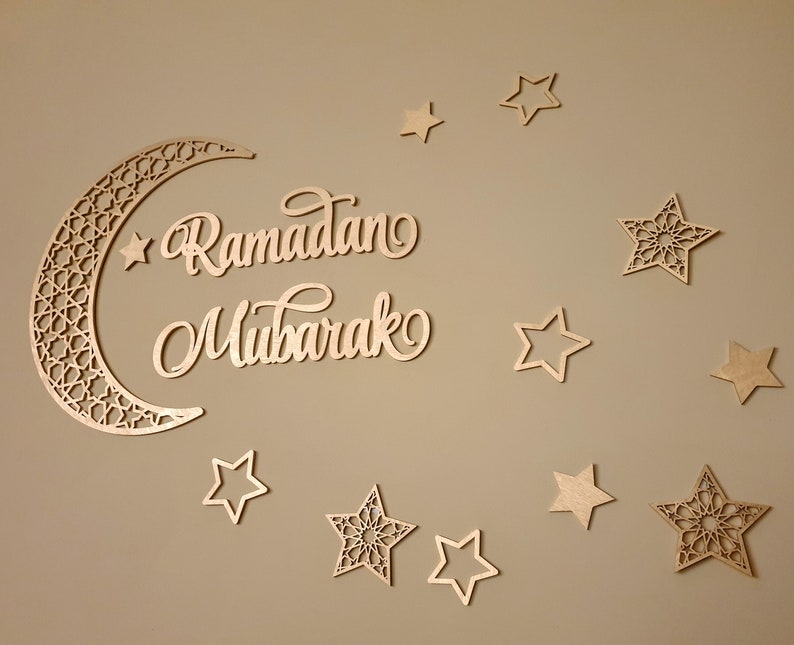 May include: A wooden crescent moon with intricate cutouts and the words "Ramadan Mubarak" in cursive script. The moon is surrounded by 10 wooden stars, some with intricate cutouts and some with simple designs.
