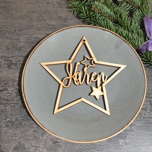 Puede incluir: Un adorno de madera en forma de estrella con el nombre "Karen" cortado en escritura cursiva. El adorno tiene una estrella m&aacute;s peque&ntilde;a en el interior y se coloca sobre un plato gris. Una ramita de hoja perenne y una cinta morada est&aacute;n en el fondo.