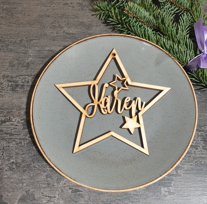 Puede incluir: Un adorno de madera en forma de estrella con el nombre "Karen" cortado en escritura cursiva. El adorno tiene una estrella m&aacute;s peque&ntilde;a en el interior y se coloca sobre un plato gris. Una ramita de hoja perenne y una cinta morada est&aacute;n en el fondo.
