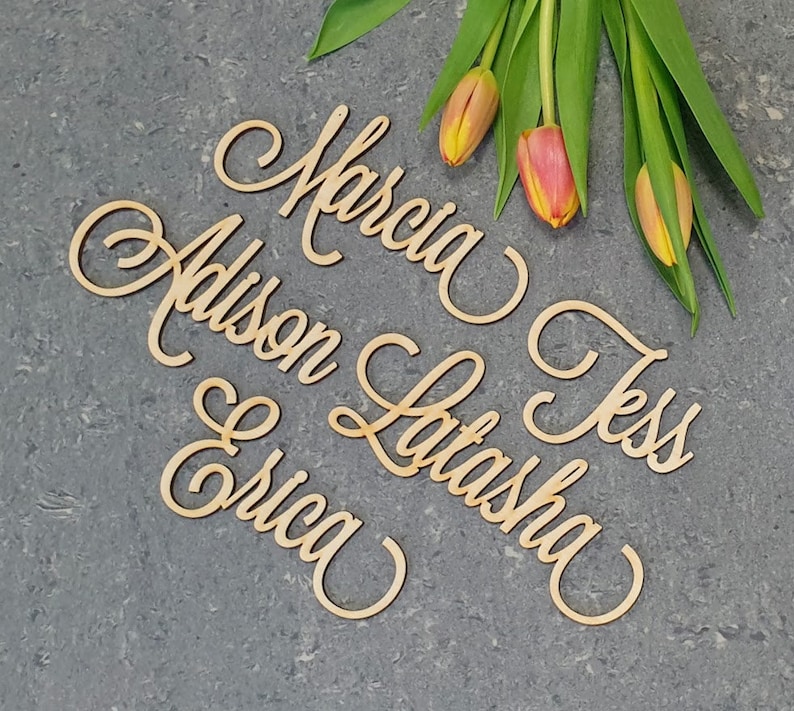 May include: Wooden cutout names in cursive script, including Marcia, Addison, Latasha, Tess, and Erica. The names are arranged in a semi-circle on a gray textured surface.  A bouquet of yellow and orange tulips is in the upper right corner of the image.