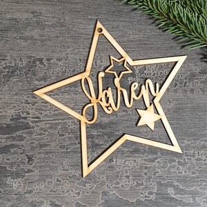 Puede incluir: Adorno navide&ntilde;o de madera en forma de estrella con el nombre "Karen" cortado en cursiva. El adorno tiene una estrella m&aacute;s peque&ntilde;a dentro de la m&aacute;s grande y un agujero para colgarlo. Una ramita de hoja perenne est&aacute; en el fondo.