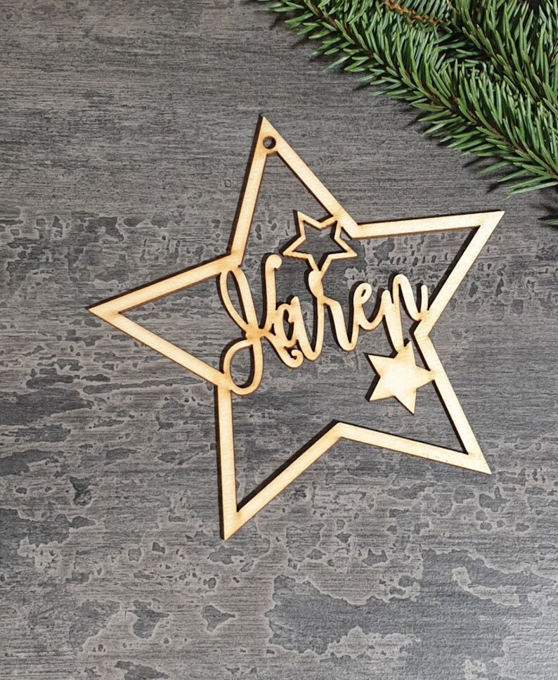 Puede incluir: Adorno navide&ntilde;o de madera en forma de estrella con el nombre "Karen" cortado en cursiva. El adorno tiene una estrella m&aacute;s peque&ntilde;a dentro de la m&aacute;s grande y un agujero para colgarlo. Una ramita de hoja perenne est&aacute; en el fondo.