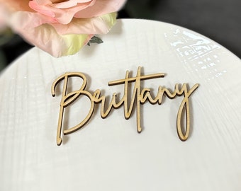 Puede incluir: Una tarjeta de lugar de madera con el nombre "Brittany" en fuente cursiva. La tarjeta es de color marrón claro y está sobre un plato blanco, con una flor rosa en el fondo. Ideal para bodas o eventos especiales.
